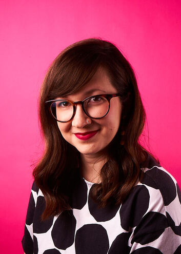 A white, queer, crip, woman with wavy brown shoulder length hair, red lipstick, glasses and a silver nose ring. She is wearing a white shirt with large black circles and smiling in front of a hot pink background.